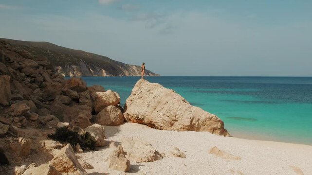 Woman standing on large rock overlooking turquoise Ionian sea waters on Fteri beach in Kefalonia Greece