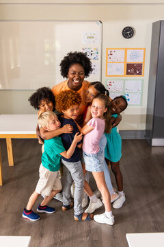 African American female teacher hugging seven schoolchildren in classroom by whiteboard, orange top
