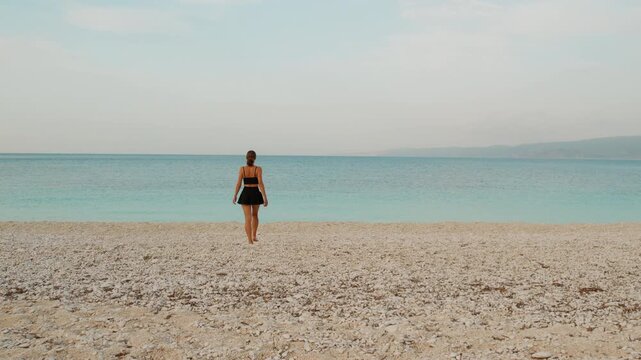 Woman walking toward calm blue sea on Kefalonia beach during serene summer day