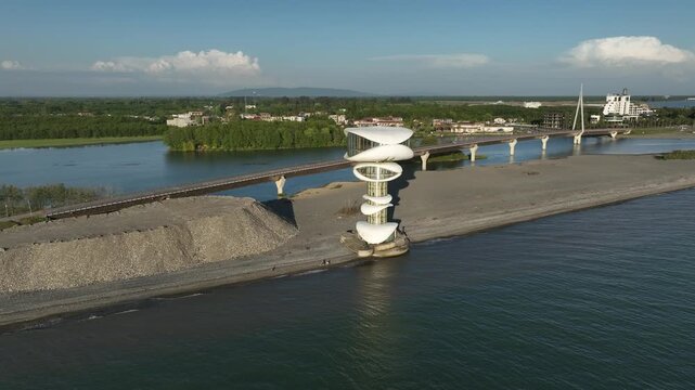 Aerial view of Anaklia resort in Georgia featuring the futuristic observation tower, pedestrian bridge over Enguri river, sandy beach, and Black Sea coastline on a sunny day.