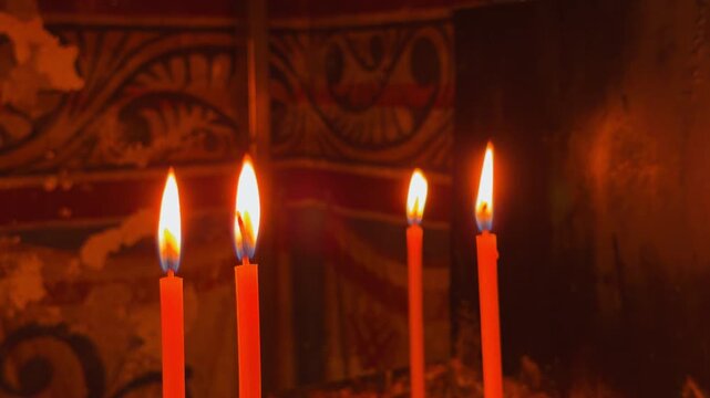 Four orange wax candle burning brightly with flickering flame in front of ornate orthodox wooden iconostasis carving inside dim church interior