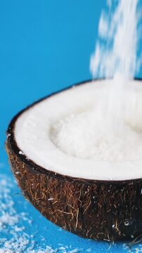 close up of white grated coconut flakes pouring into a fresh coconut half shell, tropical ingredient dynamic motion on blue background