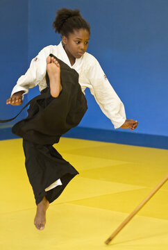 Youthful martial artist jumping to evade wooden staff strike in dojo. Dynamic aikido practice on yellow mat, cinematic portrait of agility and focus.
