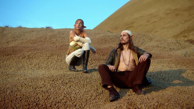 Stylish fantasy couple with elf ears posing together in a desert landscape. Cinematic lighting, flowing outfits, and surreal atmosphere create an epic fantasy-inspired fashion scene.