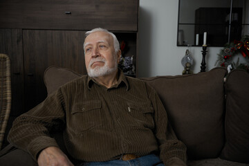 Fototapeta premium Old man sitting on couch in living room during afternoon with relaxed expression and thoughtful gaze