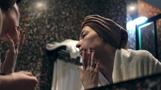 Young Caucasian woman checking her face in a bathroom mirror, looking anxious about skin condition. She gently touches her face with fingers noticing wrinkles. Skincare concern during daily routine