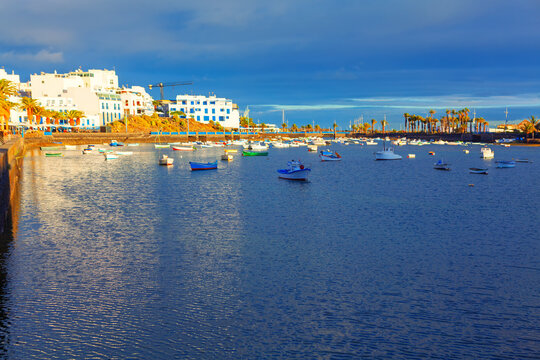 El Charco de San Gines lagoon in Arrecife, Lanzarote features rows of traditional white and blue buildings under a dramatic sky. Numerous small fishing boats bob peacefully on calm water during sunset