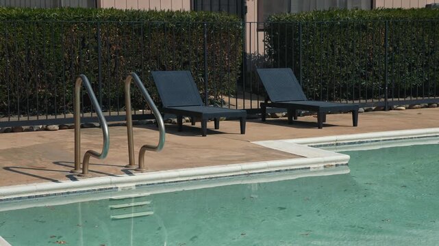 Two black lounge chairs and metal handrails by the swimming pool with clear water and dry leaves on sunny patio