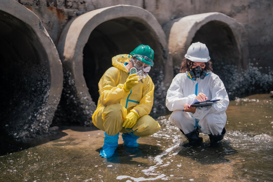 Environmental scientist documents pollution analysis results while hazmat specialist examines wastewater sample near industrial drainage infrastructure and coastal contamination site.