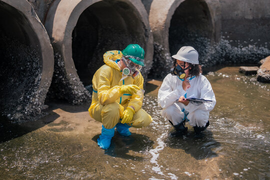 Protective suit environmental scientist documents pollution analysis results while hazmat specialist examines wastewater sample near industrial drainage infrastructure and coastal contamination site.