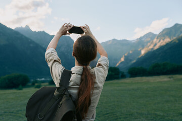 Fototapeta premium Woman with backpack taking a photo in a mountainous landscape, exploring outdoors. Bright natural light highlights nature, travel, and everyday wellness moments.