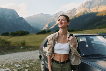 Fototapeta premium A smiling woman stands beside a SUV in a valley with towering mountains, wearing a white crop top and light jacket. A relaxed travel moment showcasing outdoor lifestyle.