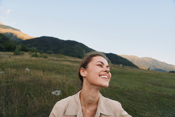 Fototapeta premium A woman in a light jacket smiles in a sunlit meadow, with rolling hills in the background, capturing a candid outdoor lifestyle moment of happiness.