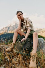 Fototapeta premium Young woman sits on a rocky mountainside with a backpack and boots, wearing khaki shirt, as sunlit meadow and distant peaks frame her thoughtful, relaxed moment