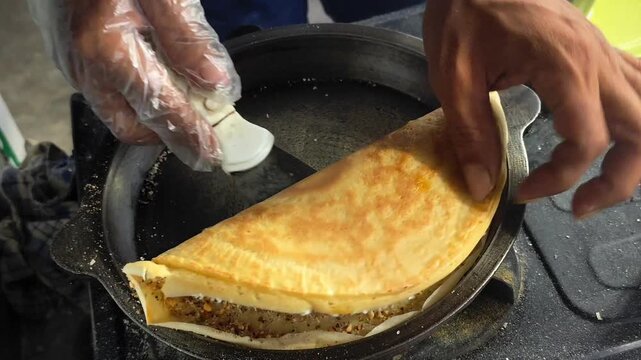 Close up footage of hands folding a traditional Indonesian sweet pancake in a cast iron pan during food preparation, showing the cooking process, peanut filling, golden texture, and authentic street f