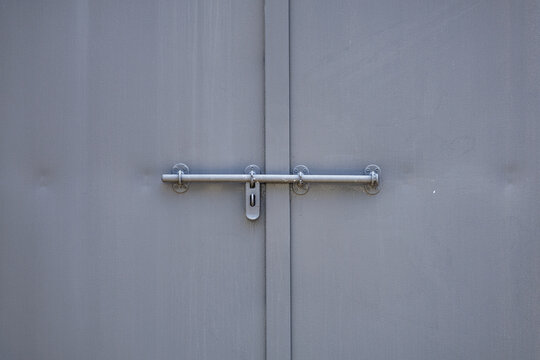 Twin muted‑gray industrial doors with padlock hasp mounted on single gray slide bolt. Minimalist industrial design emphasizing utilitarianism and security.