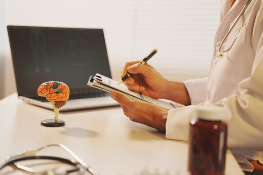 A focused shot of a doctor&rsquo;s hand writing on a clipboard with a chart. A miniature brain model stands on the desk, near a laptop showing a patient outline graphic.  