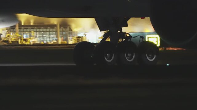 Dramatic low-angle close-up of a wide-body aircraft main landing gear rolling on the runway at night, six-wheel bogie in sharp detail with bokeh terminal lights glowing in the background