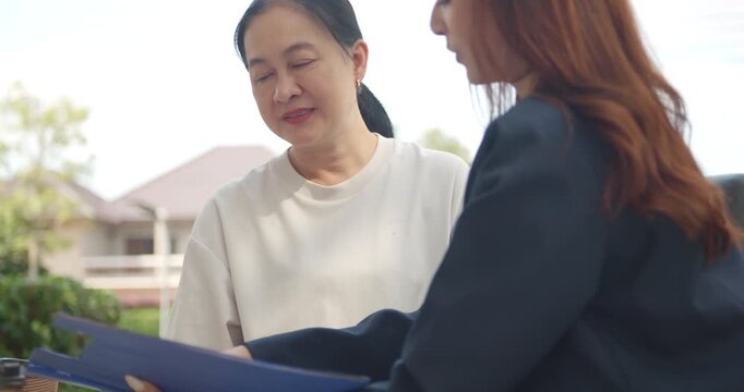 Senior Asian woman smiling and looking at documents with a female agent or financial consultant outdoors.