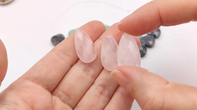 Hands hold and position three translucent light pink rose quartz briolette crystal beads with detailed geometric facets arranged next to colorful loose gemstone supplies on a white desk
