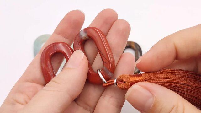 Hands inspect a polished red jasper oval chain link element featuring a rich terracotta hue with natural veining attached to a matching silky brown decorative textile tassel accessory