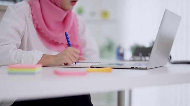 Muslim businesswoman entrepreneur writing notes planning work strategy using laptop computer at modern office desk workspace professional female wearing hijab