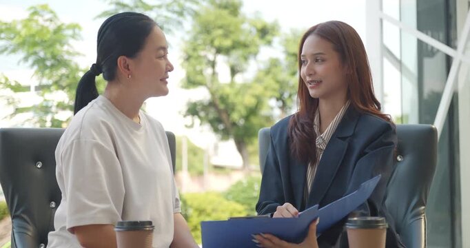Young Asian businesswoman presenting financial or insurance documents to a mature female client at an outdoor cafe.