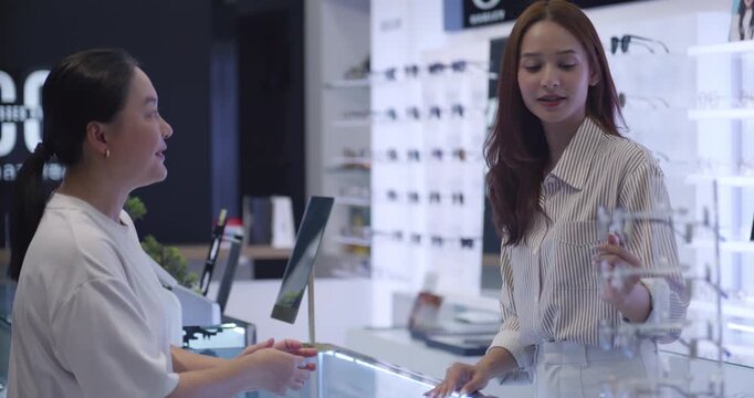 Senior Asian woman trying on new eyeglasses with the assistance of a smiling shopkeeper in an optical store.