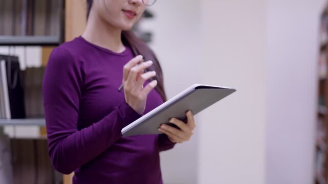young Asian women librarian office worker professional hold tablet digital device check inventory standing in library archive bookshelf background workspace