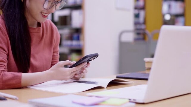 Asian student using smartphone mobile phone studying with laptop computer textbook at library desk for online learning education research browsing social media
