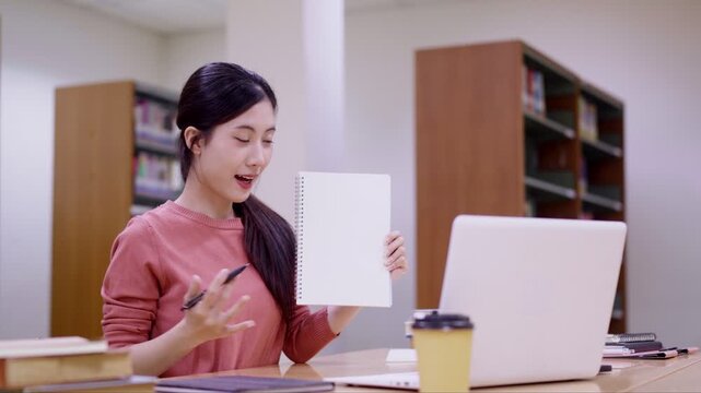 Asian student women holding notebook writing pen study research education learning library workspace using laptop computer reading book indoor bright room