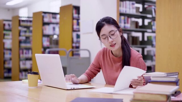 Asian female student using laptop computer researching studying at university library reading notes doing online learning homework assignment with books coffee