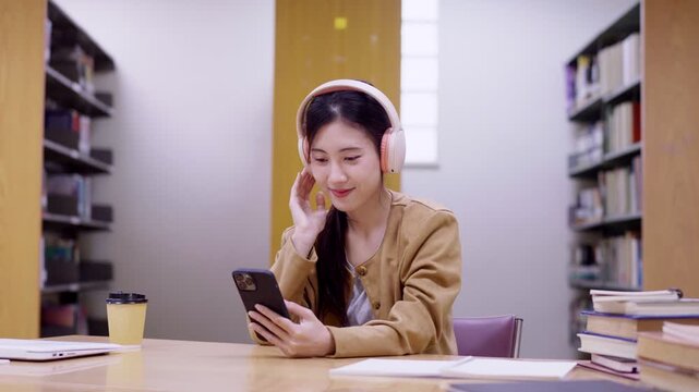 young Asian student women using smartphone with headphone listening music podcast online streaming at library study room with book stack learning knowledge