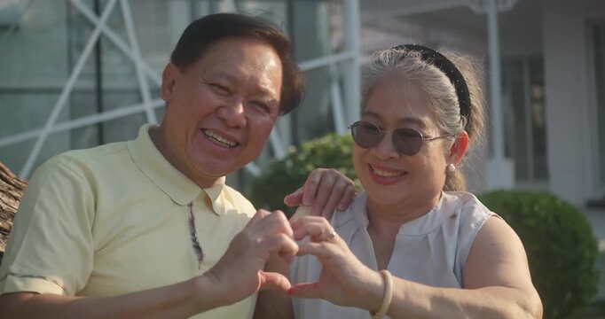 Happy retired senior Asian couple making a heart shape with their hands and smiling at the camera outdoors.