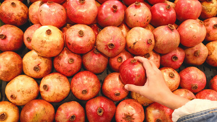 Hand Selecting a Fresh Red Pomegranate from a Vibrant Market Display © blueringmedia