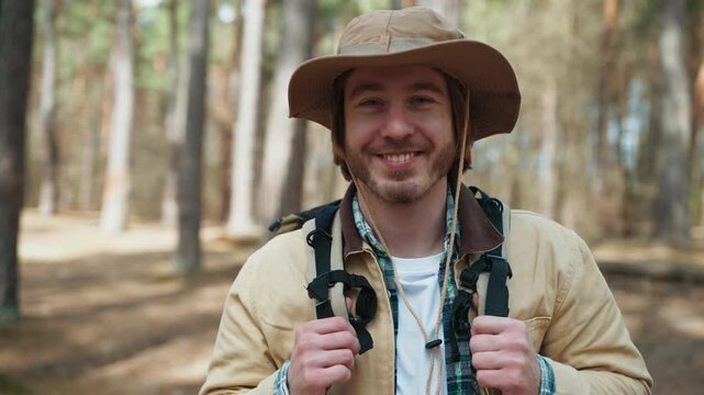 Smiling Caucasian backpacker standing in sunny woodland. Handsome male traveler wearing hiking hat and carrying gear outdoors. Tourist enjoying freedom and active lifestyle during forest expedition.