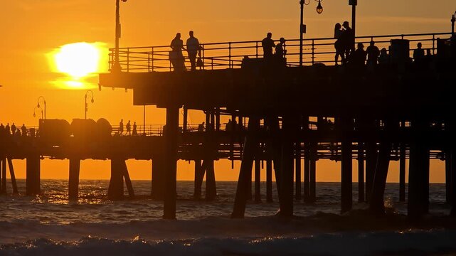 Santa Monica Pier at sunset with silhouetted visitors over Pacific Ocean waves, glowing orange sky and iconic coastal structure in California