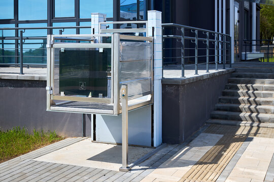 Modern vertical platform lift provides barrier-free accessibility to building entrance. Metallic structure features safety rails and non-slip surface, ensuring secure outdoor mobility for users.