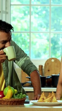 Indian asian young husband and wife having coffee in modern kitchen platform with light hearted chit chat enjoying their morning beverage and talking together in a relaxed home environment