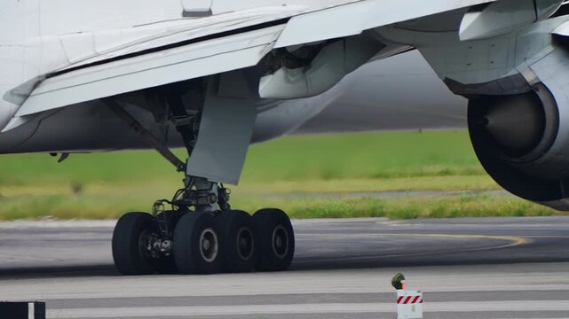 Close-up of a wide-body commercial aircraft's main landing gear six-wheel bogie rolling on the runway, with the wing underside and deployed flaps visible overhead