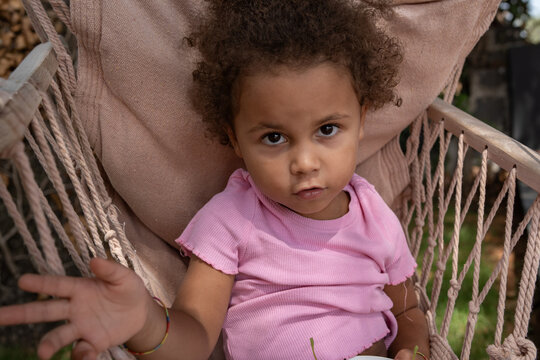 Cute mixed race toddler girl relaxing on hanging chair in summer garden
