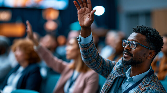 Male professional raising his hand to pose a question during an interactive business seminar in a large conference hall, other attendees visible turning to look, defocused presenter and