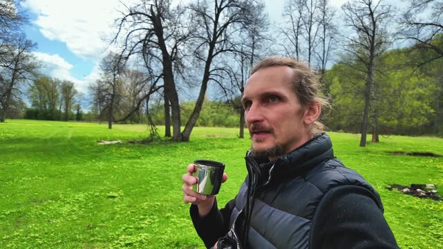 Caucasian man tasting coffee on trail during travel vlog walk, animated profile shot in open meadow with tall bare trees and bright green grass, cloudy sky, handheld camera, casual puffer