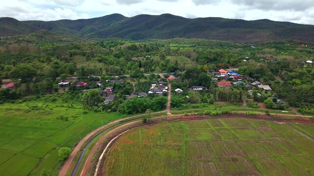 Beautiful drone footage showing a scenic tropical village nestled between lush green agricultural fields and majestic mountain ranges under a cloudy sky Daytime aerial landscape in Thailand