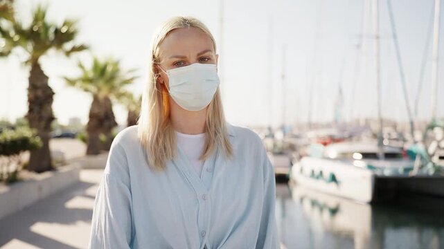 Blonde woman wearing protective face mask walking at marina with yachts on a sunny day. Travel safety, health precautions, and modern travel lifestyle concept.
