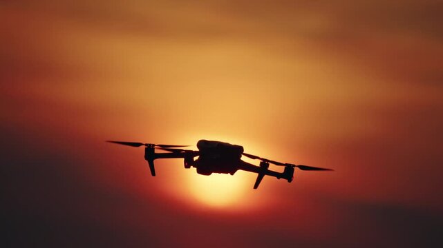 A drone hovering and slowly descends against a vibrant orange and red sunset sky in a cinematic outdoor shot.
