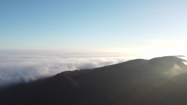 Drone footage of dark mountain peaks emerging from a thick white sea of clouds. Camera captures a bright horizon with soft sunlight shining over the foggy inversion layer.