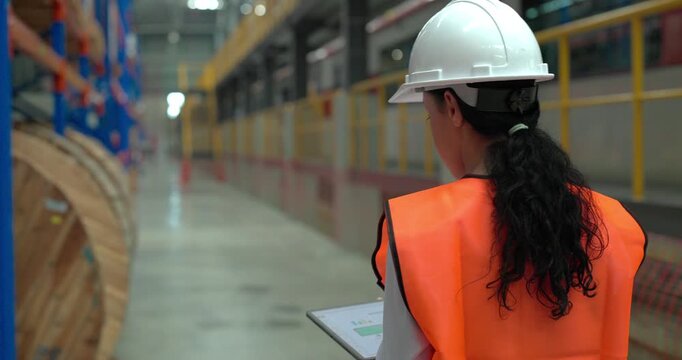A female rail engineer wearing a hard hat and reflective vest inspects inventory, scans shelves, and reviews checklists in a large railway supply warehouse depot
