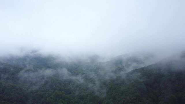 Mysterious aerial view of mountain peaks disappearing into thick white fog and low storm clouds, dramatic moody nature landscape from drone.