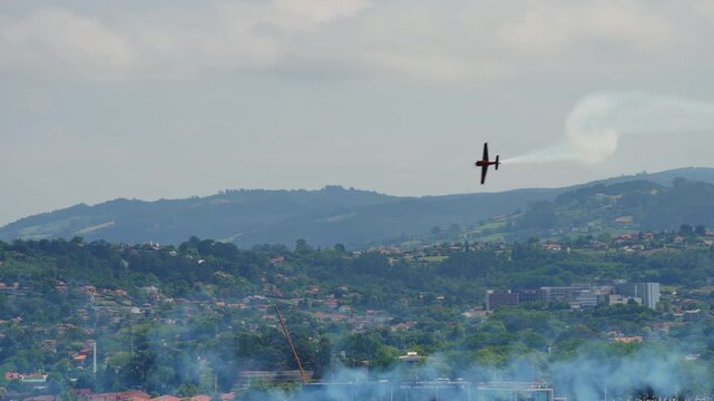 A fast aerobatic aircraft trailing white smoke performs a maneuver at an airshow above a hilly residential landscape with blue pyrotechnic smoke drifting in the foreground